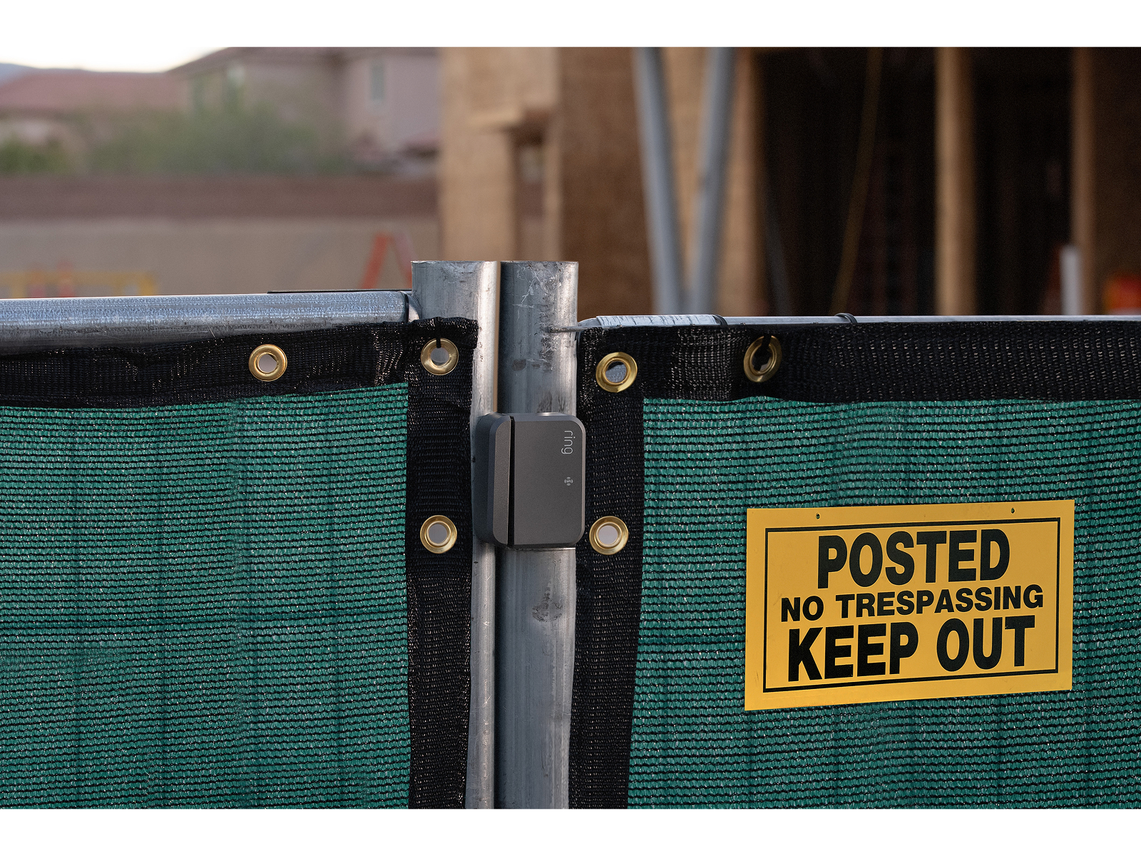 Green mesh construction fence with metal posts, a small gray Ring device on the seam, and a yellow “Posted No Trespassing Keep Out” sign.