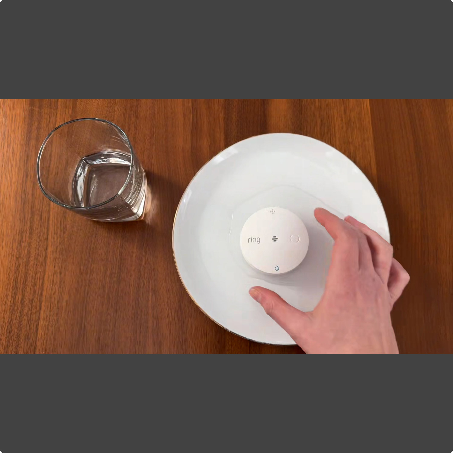 Flood & Freeze Sensor (Newest Model) - Hand reaching for a white Ring sensor on a white plate, next to a glass of water on a wooden table.