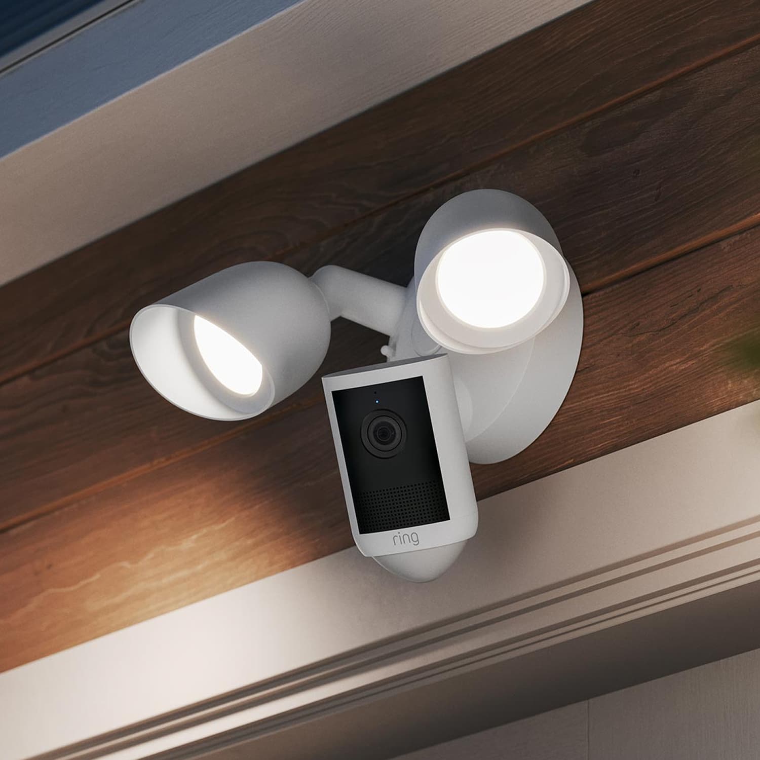 Floodlight Cam - White outdoor floodlight with integrated security camera mounted under a wooden eave, both lights turned on at dusk.