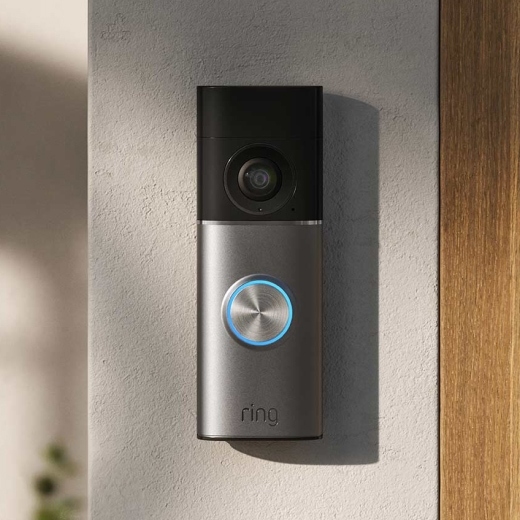 Sleek video doorbell mounted on a stucco wall, black and silver body with a glowing blue ring button and camera lens above.