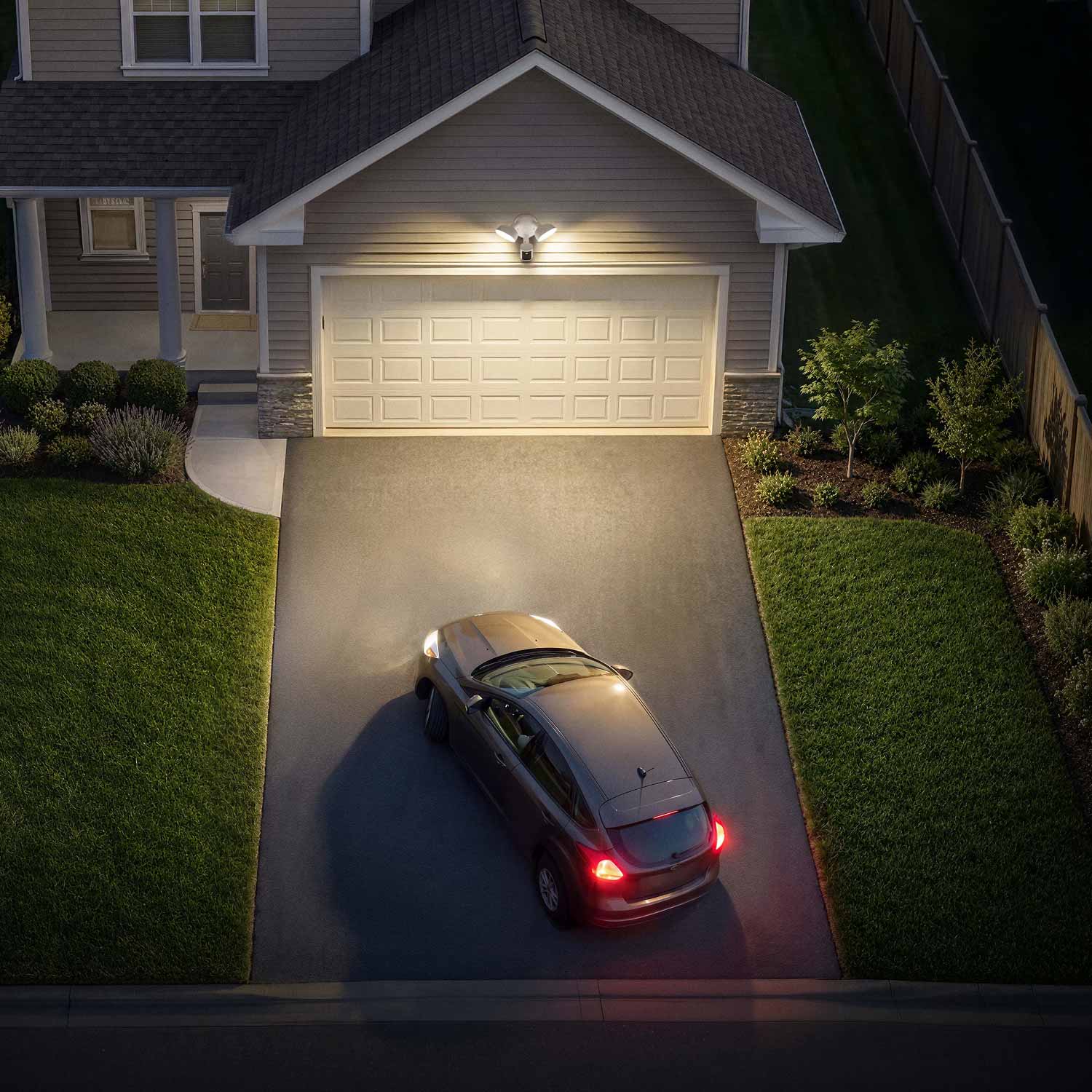 Car turning into a suburban driveway at dusk, garage lights illuminating the white door and neatly landscaped front yard.