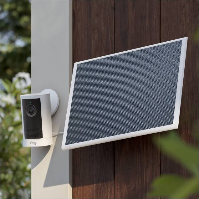 Outdoor Ring camera mounted beside a tilted solar panel on a wooden fence, with greenery softly blurred in the background.
