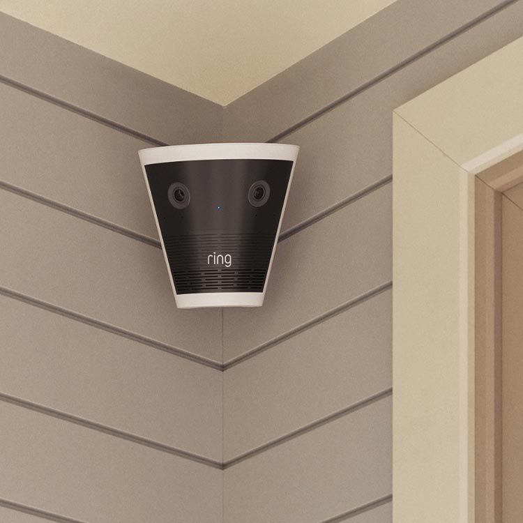 Corner-mounted Ring security camera with dual lenses and a blue status light on gray shiplap siding near a doorway.