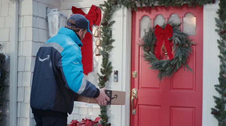 Amazon delivery person placing package at festive red door decorated with Christmas wreath and garland.