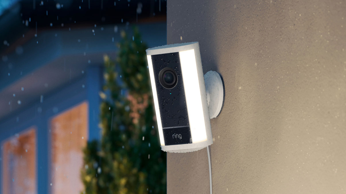 Wall-mounted Ring floodlight camera in the rain, glowing white side lights, wet lens, with a house window and shrub softly blurred in the background.