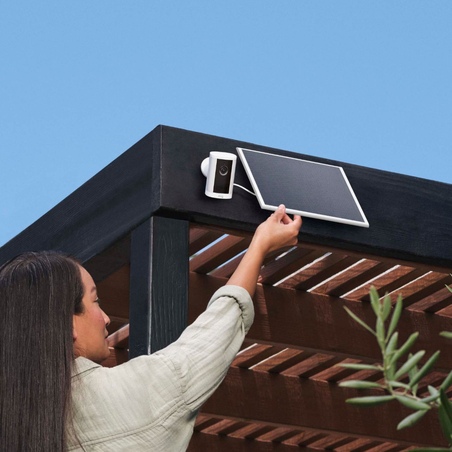 Person mounting a white solar panel beside a small outdoor camera on a dark wooden pergola under a clear blue sky.