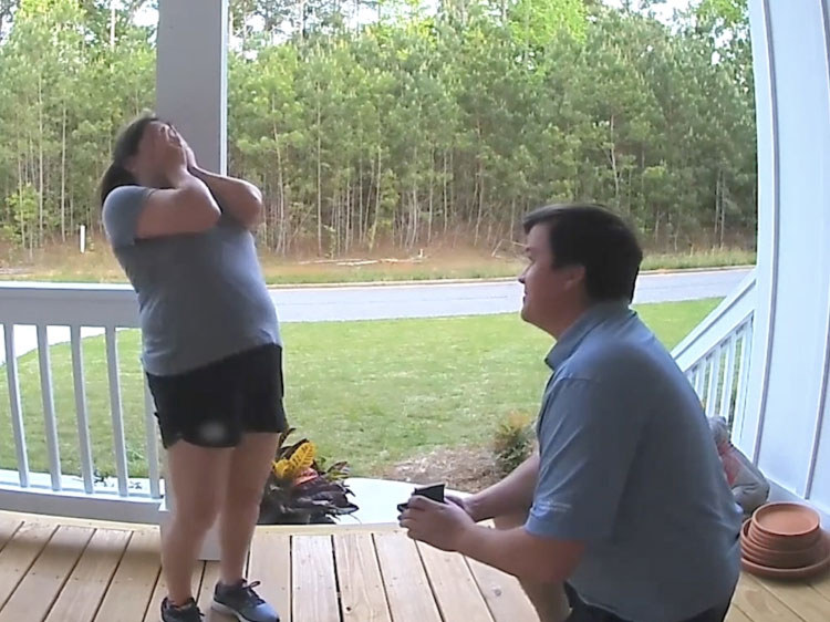 A man on one knee proposes to a surprised woman on a front porch.