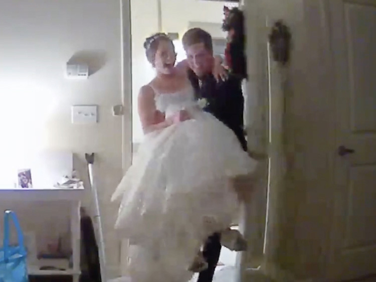 A man in a suit carries a woman in a wedding dress through an interior doorway.