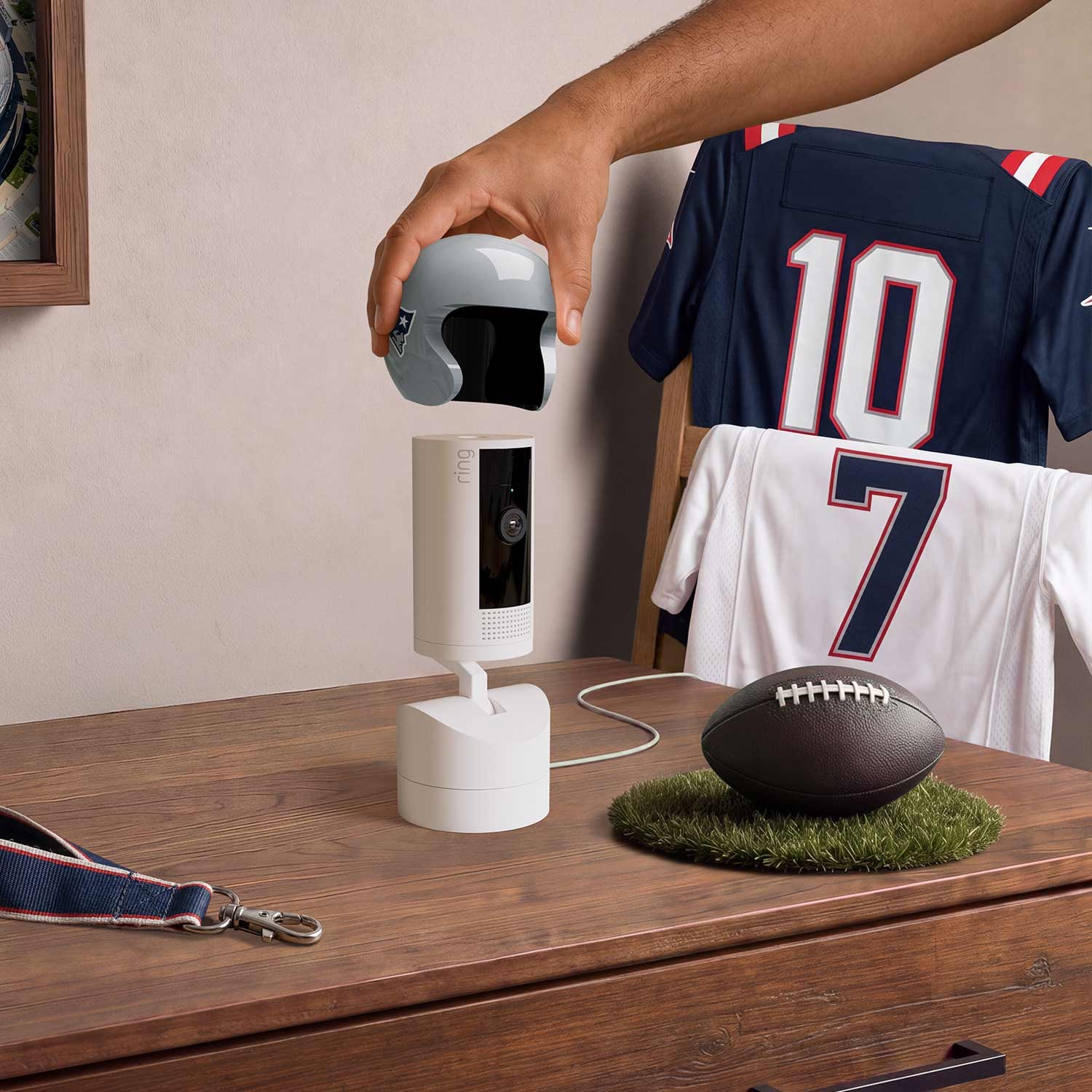 Hand placing a navy football helmet cover on a white indoor camera on a wooden table, with jerseys on a chair and a football nearby