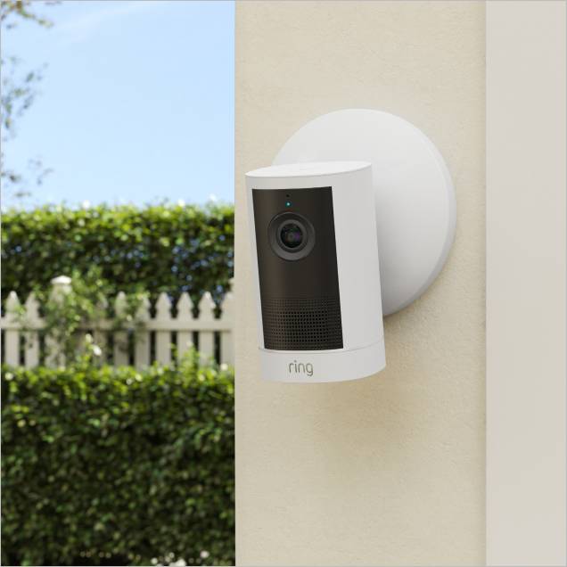 White Ring outdoor security camera mounted on a beige wall, facing a garden with a white picket fence and green hedges in daylight.