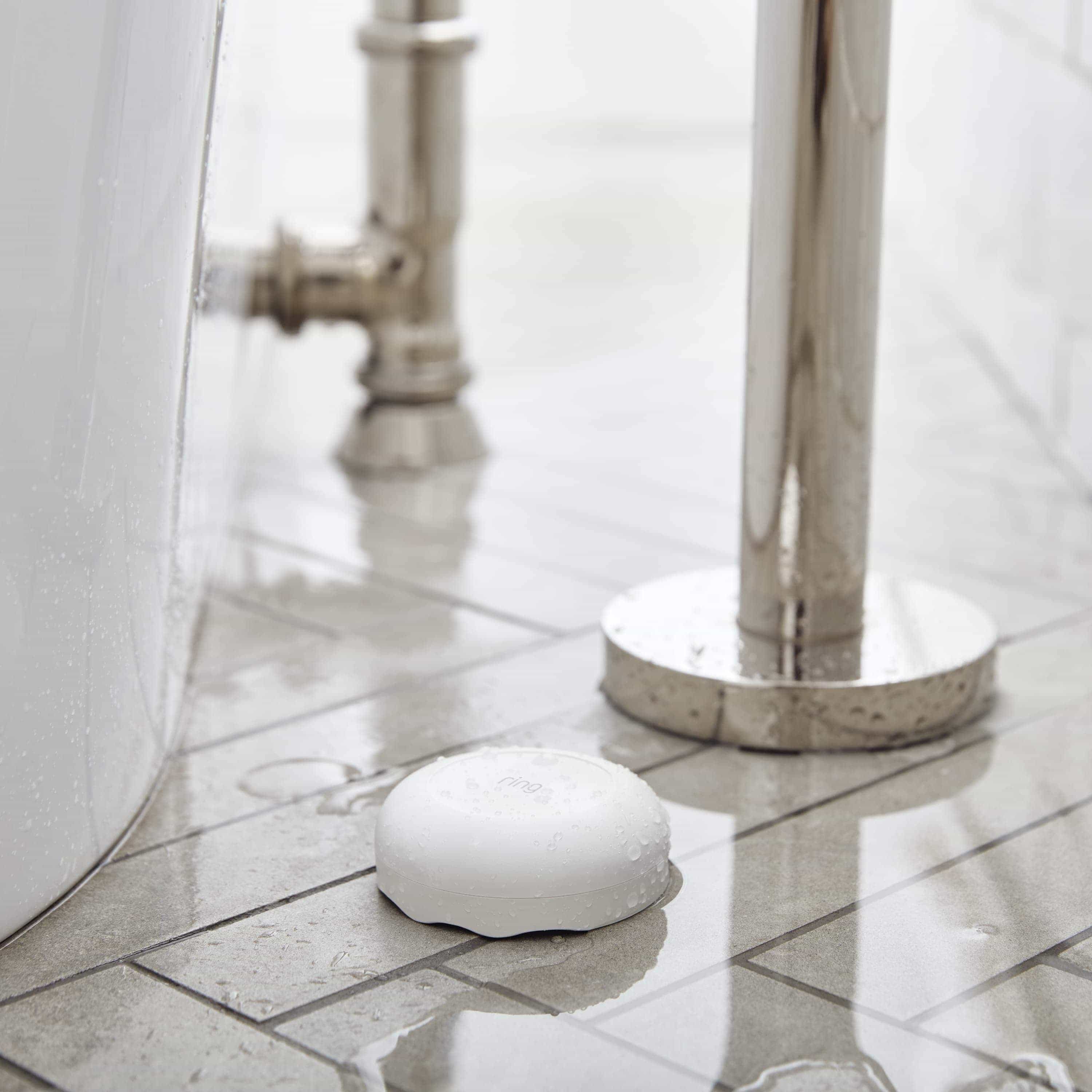 Alarm Flood & Freeze Sensor - Alarm Flood and Freeze Sensor situated on a wet, tiled floor next to a bathtub and plumbing fixtures.