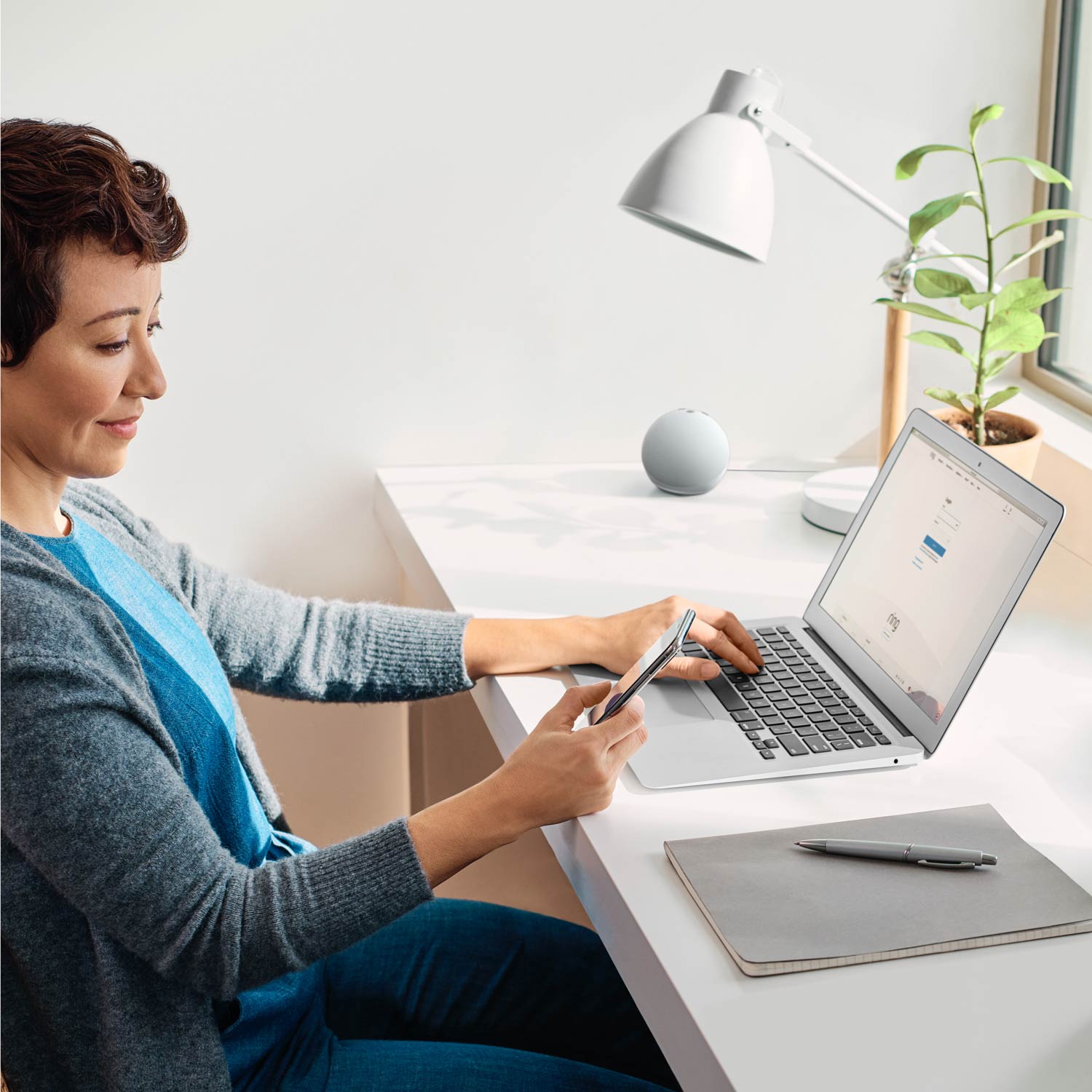 Alarm Pro Base Station (for with built-in eero Wi-Fi 6 router) - Side-view of woman sitting at desk in front of laptop with Ring web page loaded, looking at smartphone in her right hand.