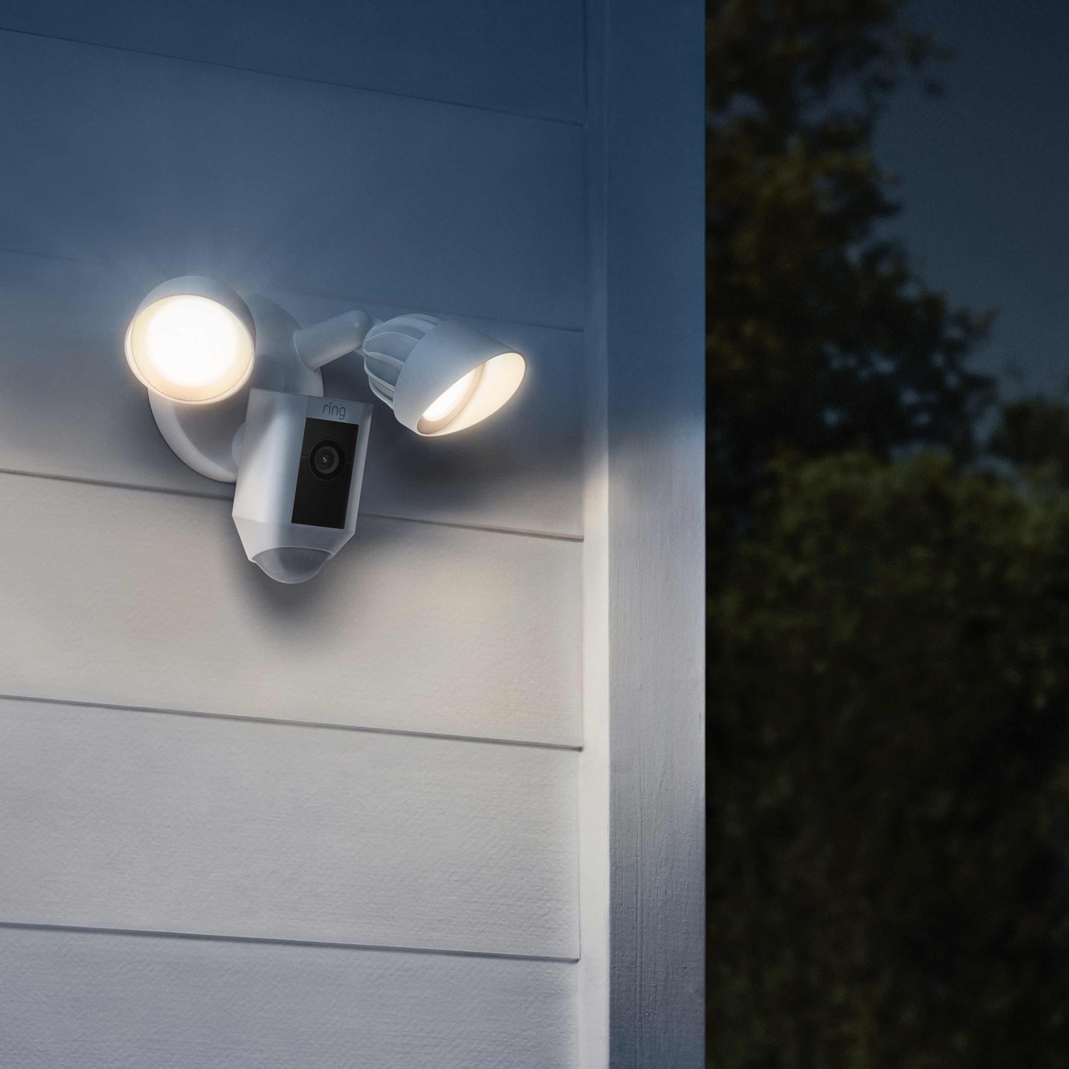 Floodlight Cam Wired Plus (Certified Refurbished) - Outdoors at night, an illuminated Floodlight Cam Wired Plus in white is mounted to an exterior wall of home near the corner.
