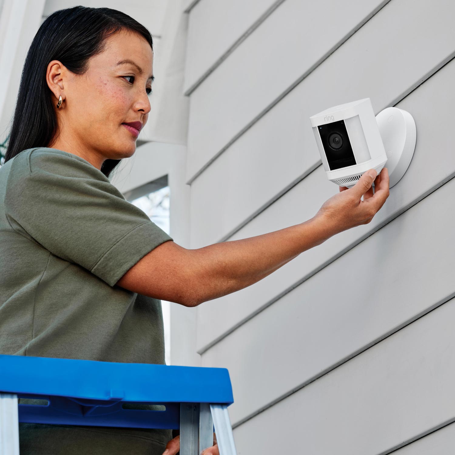 Spotlight Cam Plus (Wired) - Woman on a ladder installing Spotlight Cam Plus, Wired model in white on exterior wall of home.