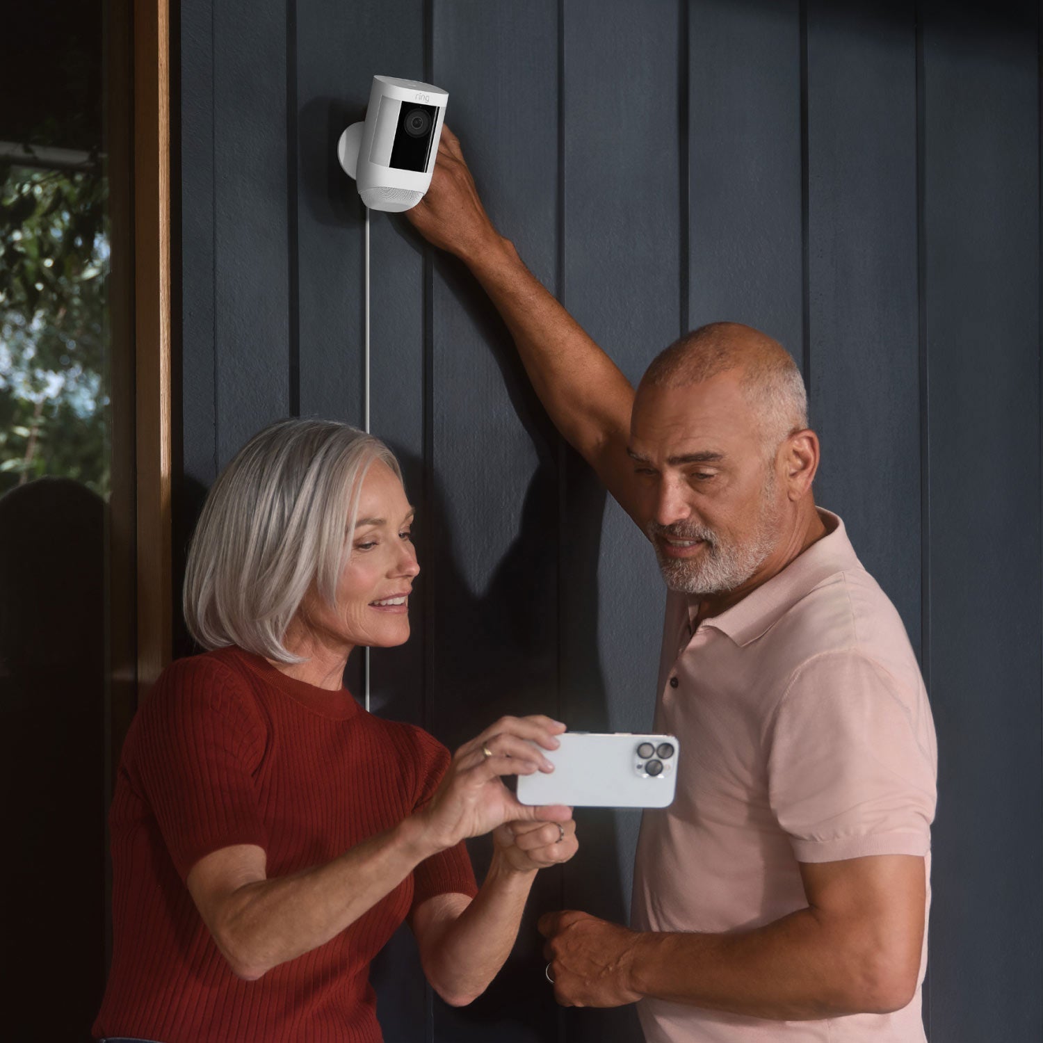 2-Pack Spotlight Cam Pro (Plug-In) - Woman and man looking at smartphone while installing Spotlight Cam Pro Plug-In model on exterior wall of home.