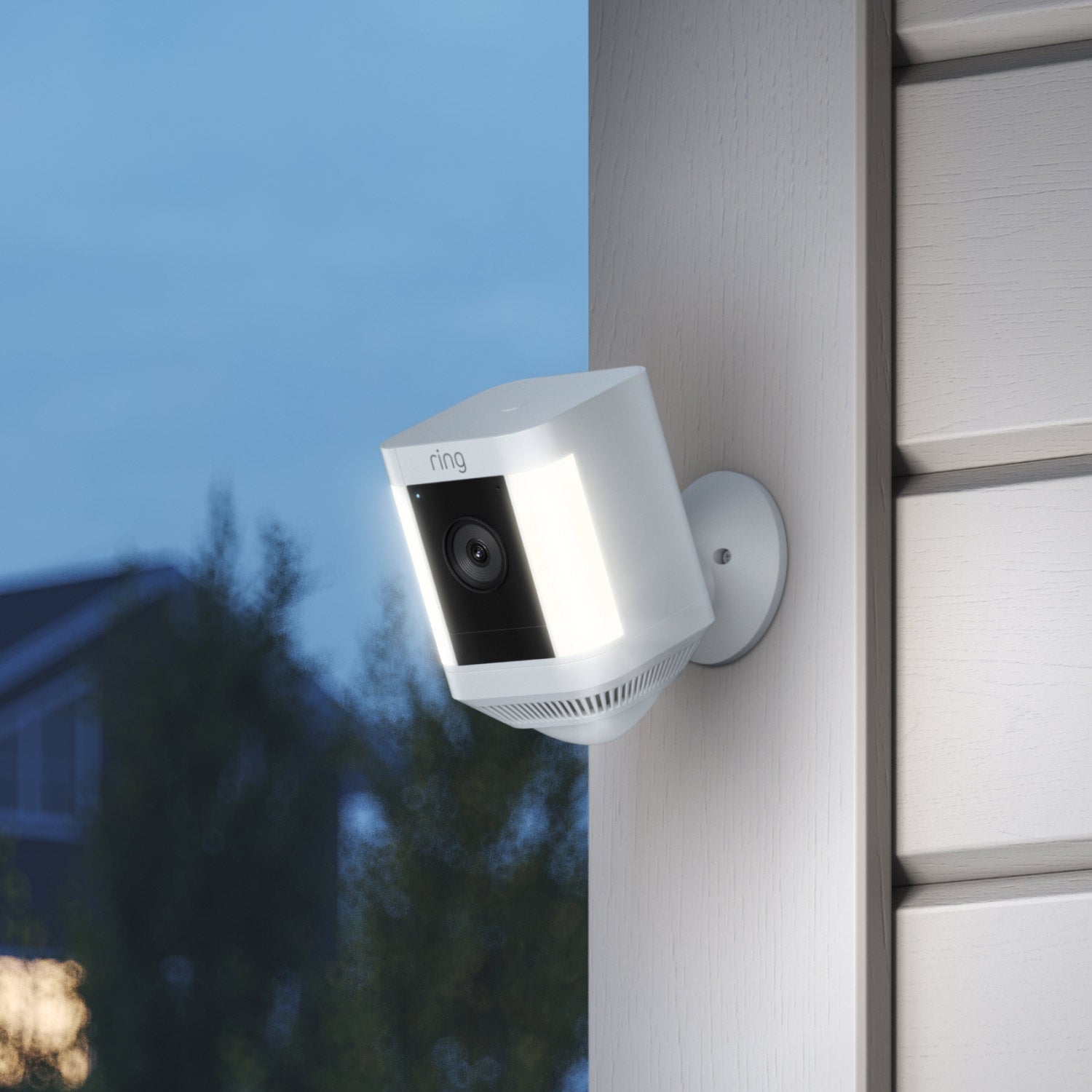 2-Pack Spotlight Cam Plus (Battery) - Outside at dusk, an illuminated Spotlight Cam Plus is mounted to a wall near the corner of a home.