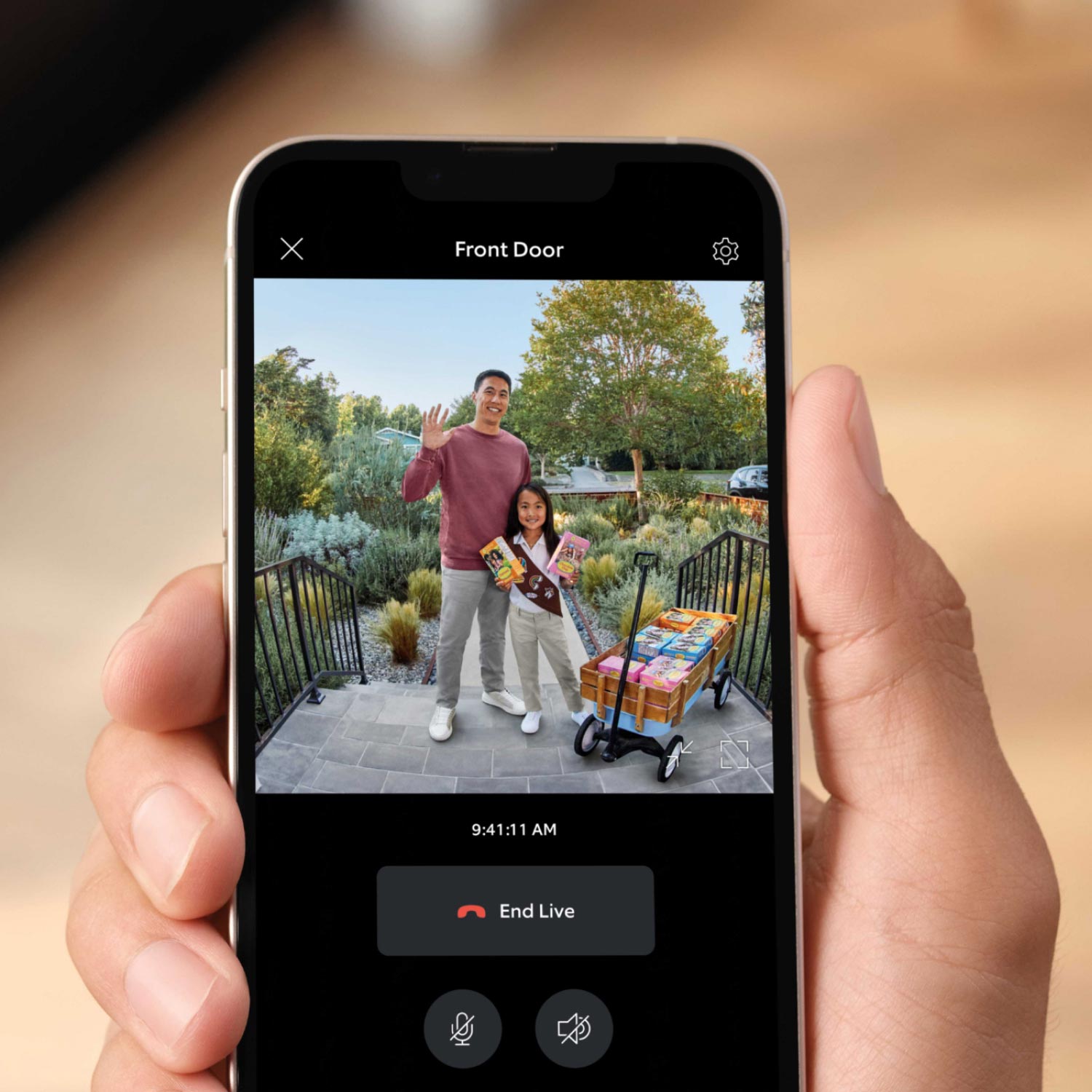 Battery Doorbell Plus - Close-up of a hand holding a smartphone. The screen shows video of a smiling man and a girl at the front door with a wagon full of items.