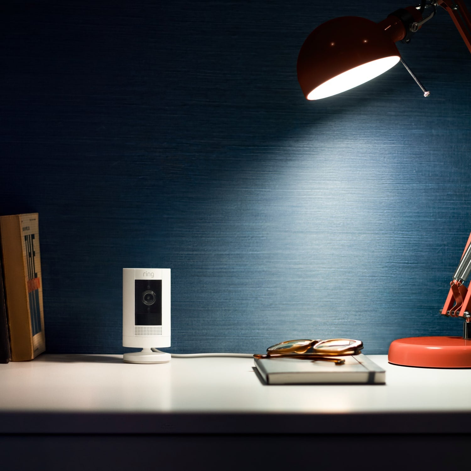 Stick Up Cam Plug-In (for Certified Refurbished) - Stick Up Cam, Plug-In model in white on a desktop next to a pair of glasses, a book, and a lamp.