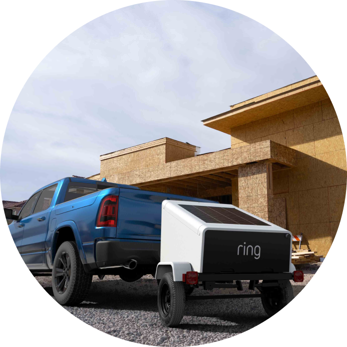 Blue pickup towing a small white Ring trailer with a solar panel at a wood-framed construction site under a cloudy sky.