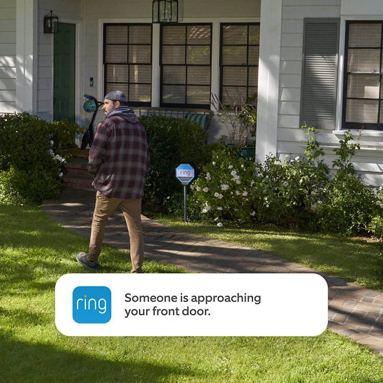 Person walking up a brick path toward a house’s front door, garden and scooter by porch, with a Ring sign in the yard.