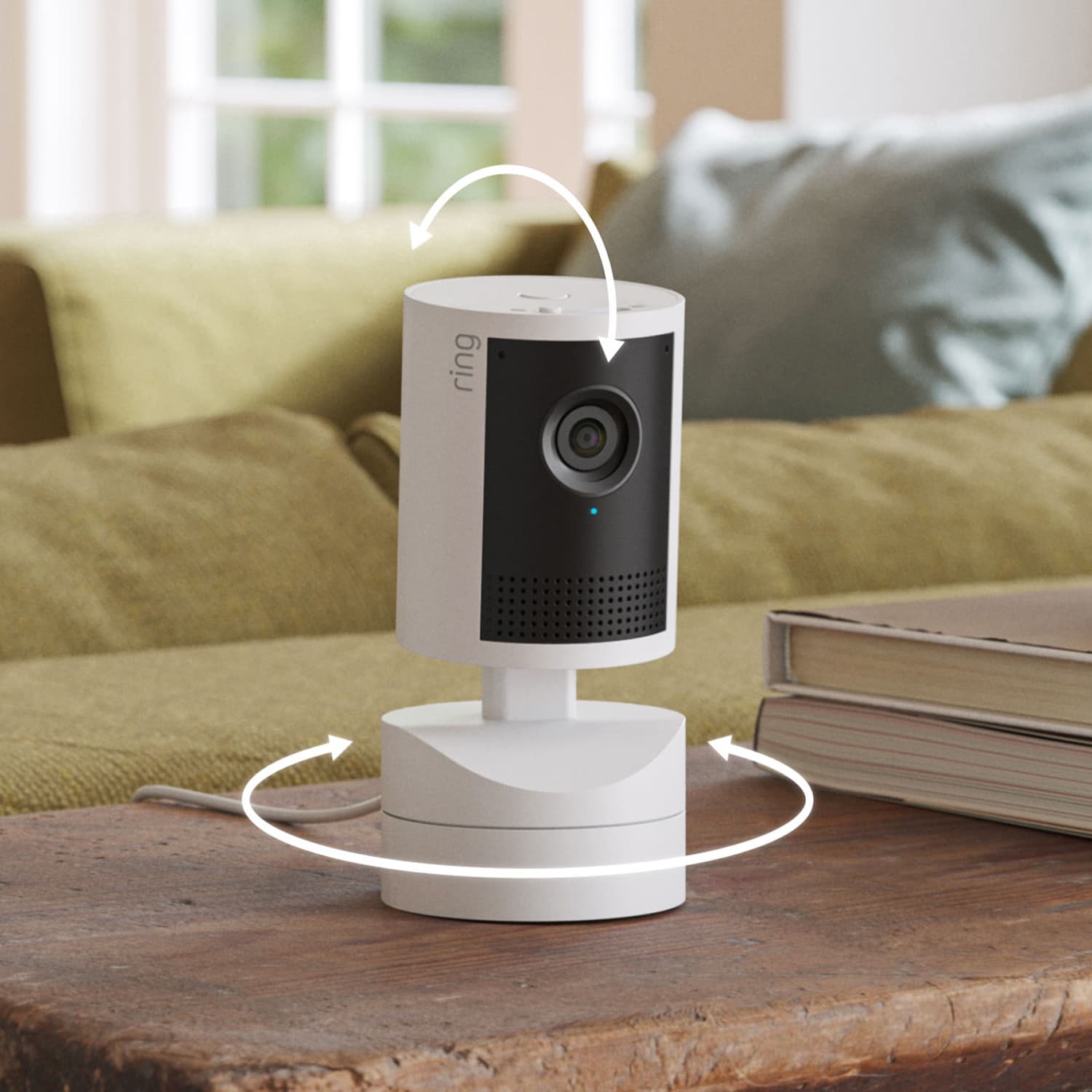 Pan-Tilt Indoor Cam 2K - White indoor security camera on a wooden table, showing tilt and pan arrows, with a sofa and stacked books in a cozy living room.