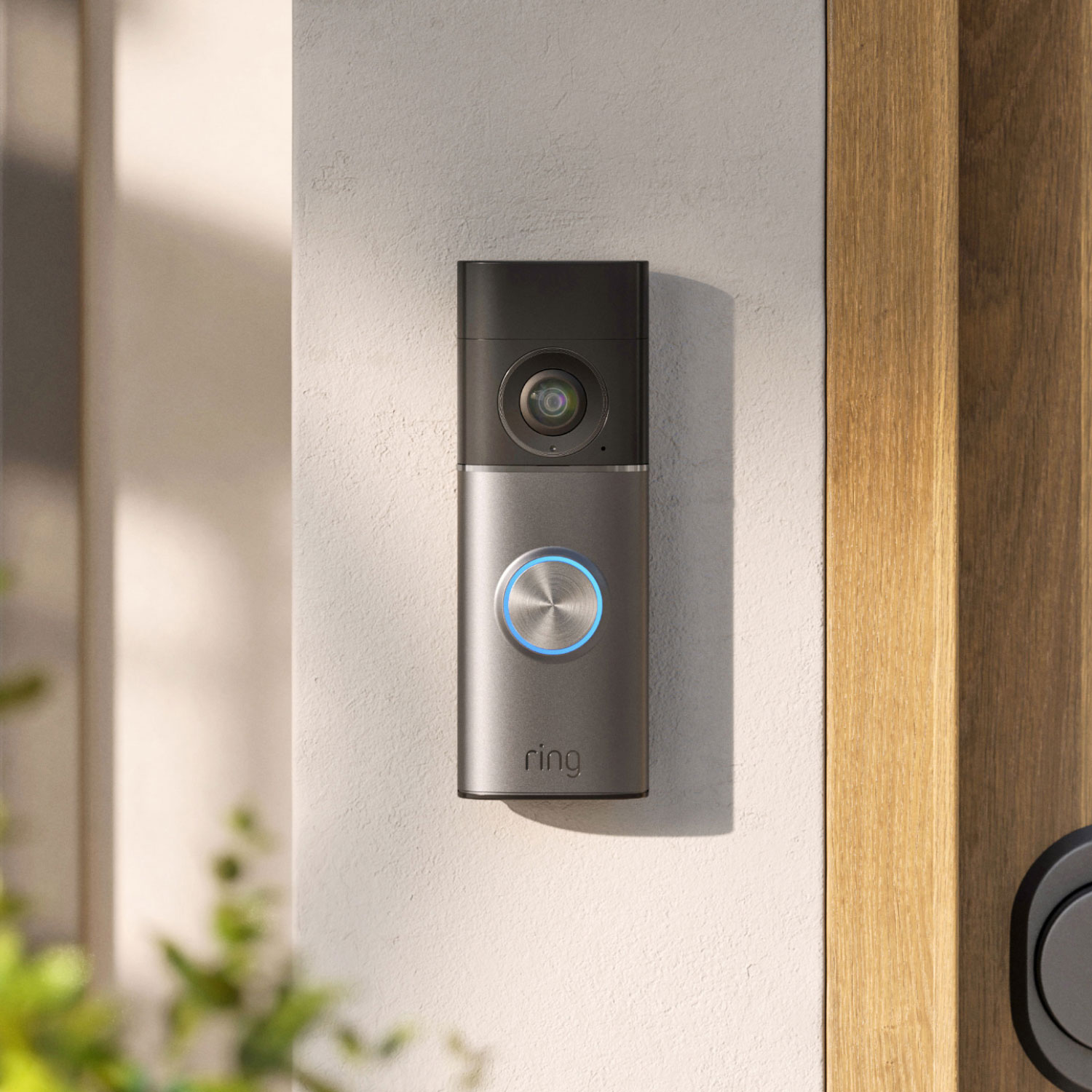 Smart video doorbell with a glowing blue ring button mounted on a light wall beside a wooden door, in soft daylight.