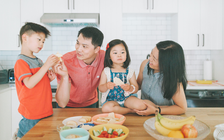A family of four with two young kids in the kitchen wearing summer clothes with fresh fruit on the counter.