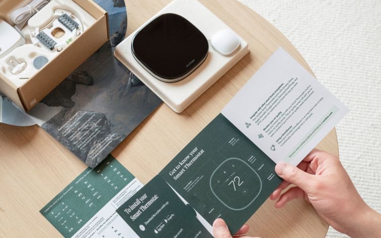 A person reading and holding the ecobee Smart Thermostat quickstart guide for installing the thermostat while the packaging and product for the thermostat rests on a table.