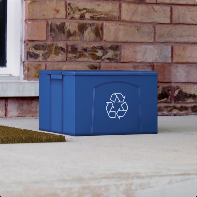 A box made to look like a recycling bin sits on a porch in front of a door against a brick wall. 