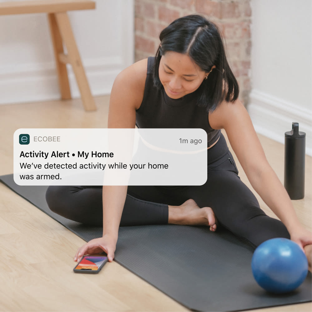 A woman looks at her phone, with an ecobee notification saying "activity alert at my home"