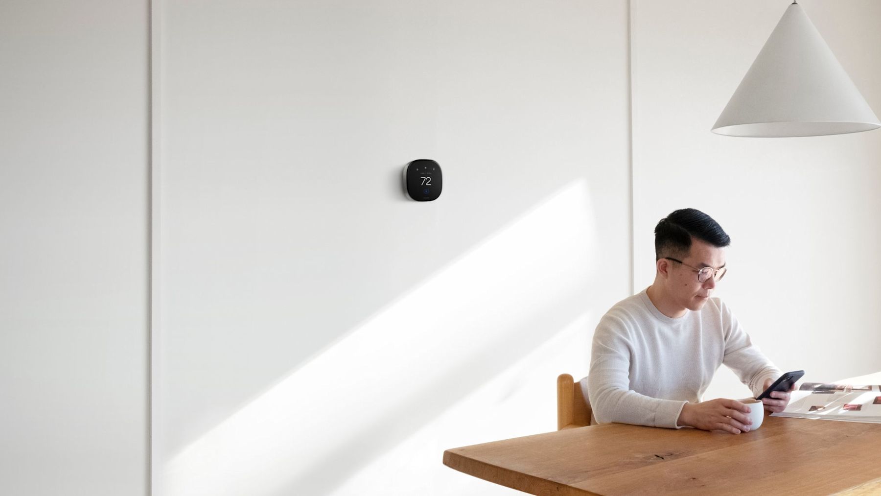 ecobee's Smart Thermostat Premium on a white wall in an home with a man sitting at a table in front of it, looking at his phone and drinking a coffee.