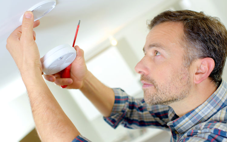 A man changes a fire alarm on a ceiling.