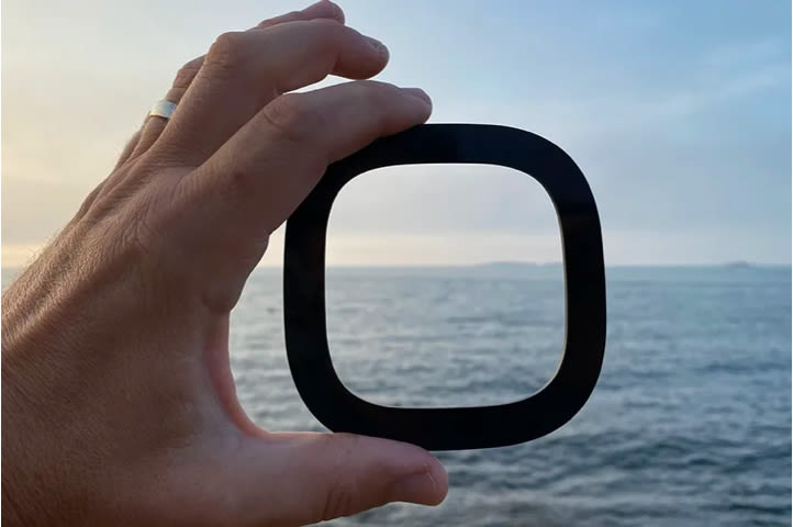 A hand holds a squircle shape in front of a body of water.
