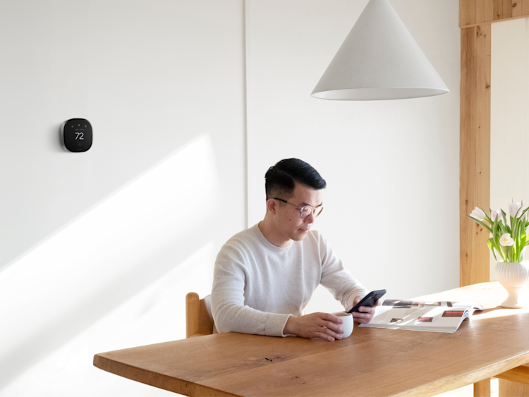 A man is sitting at his kitchen table in a contemporary home with a Smart Thermostat Premium installed on the wall in background.