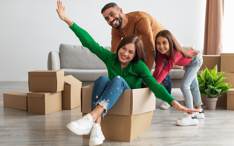 A family of three including a man, woman, and young girl, celebrate moving into a new home with moving boxes surrounding them.