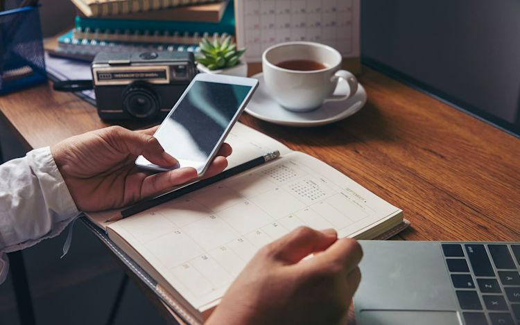 Laptop and day planner open, phone in-hand, man sips tea and contemplates the day ahead.