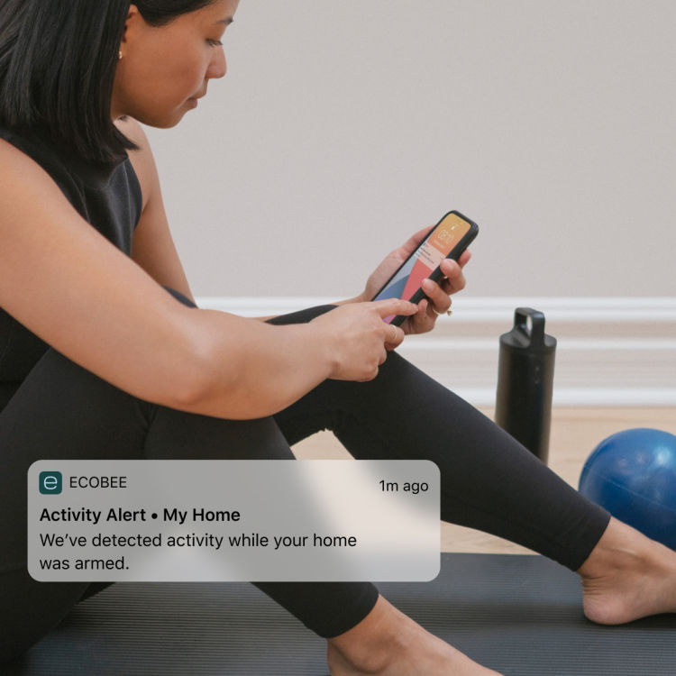 A woman checks her phone while exercising; a notification from the ecobee app says "Activity alert - My home"