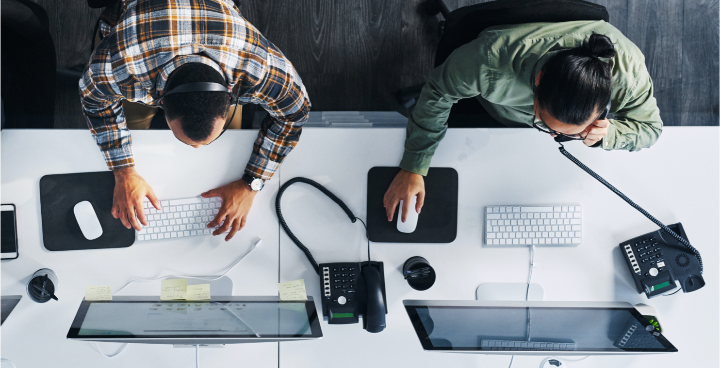 An overhead shot of two people at a desk with computers and phones