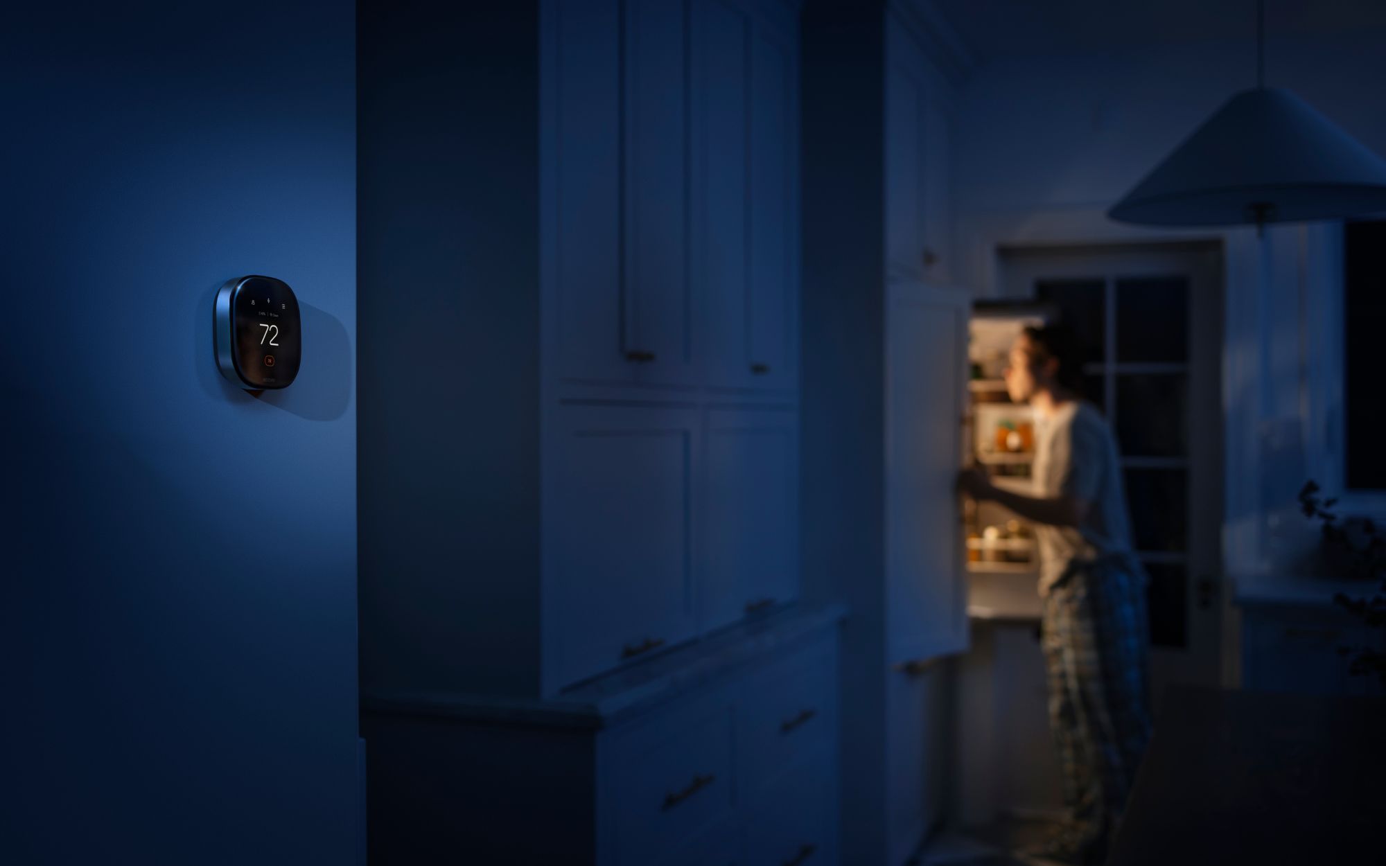 ecobee smart thermostat displaying 72°F on a kitchen wall at night as a person opens the refrigerator in the background.