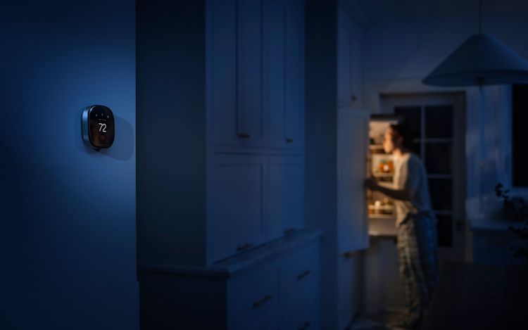 ecobee smart thermostat displaying 72°F on a kitchen wall at night as a person opens the refrigerator in the background.
