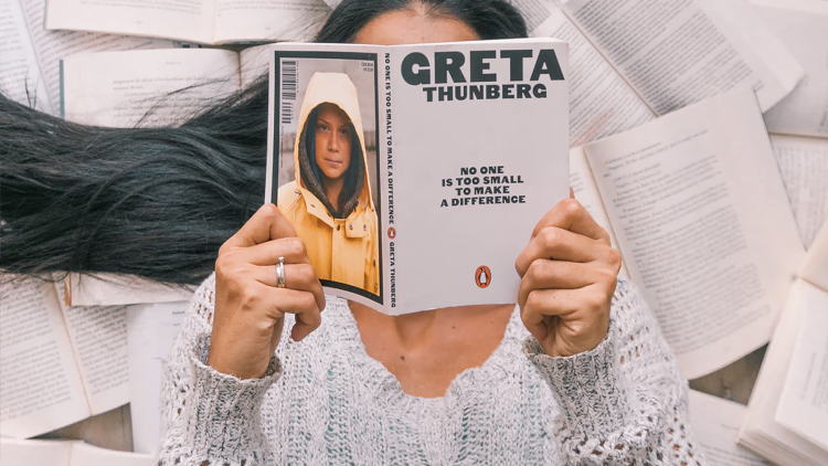 A woman laying on a pile of open books reads Greta Thunberg's "No One is Too Small to Make a Difference"