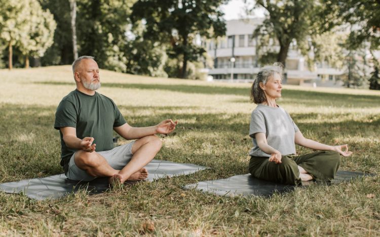 A man and a woman doing yoga in a park, in lotus position.