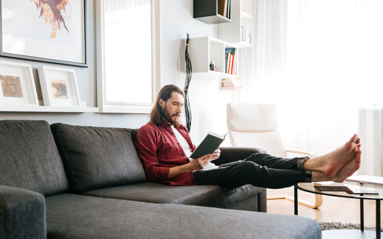 Man in unbuttoned red plaid shirt with white tee-shirt reading on couch.