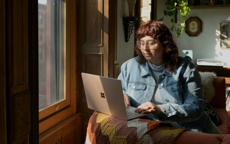 A woman sitting on a couch near the window, using her laptop.
