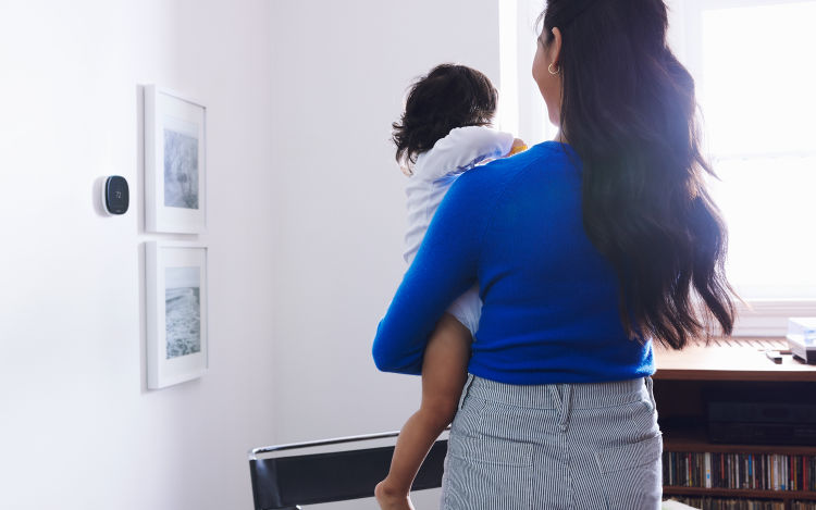 Woman in blue long-sleeve shirt adjusts temperature on her ecobee SmartThermostat with voice control handsfree while carrying toddler 