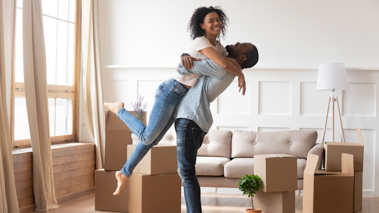 A happy couple that is a man and a woman are happy and excited about moving into a new home. The man is lifting the woman and there are moving boxes surrounding them.