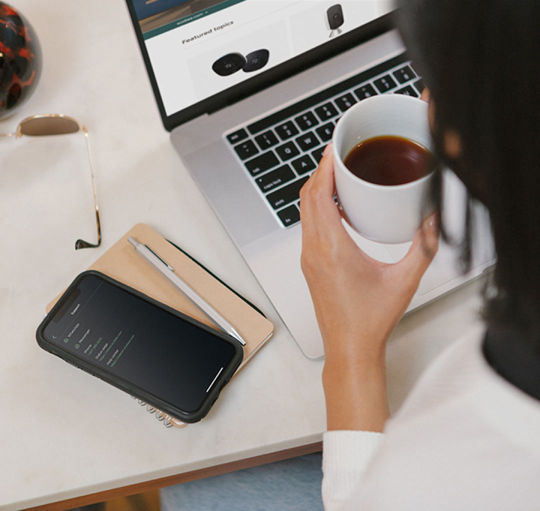 a person sitting at a laptop with a cup of coffee looks at a phone screen.