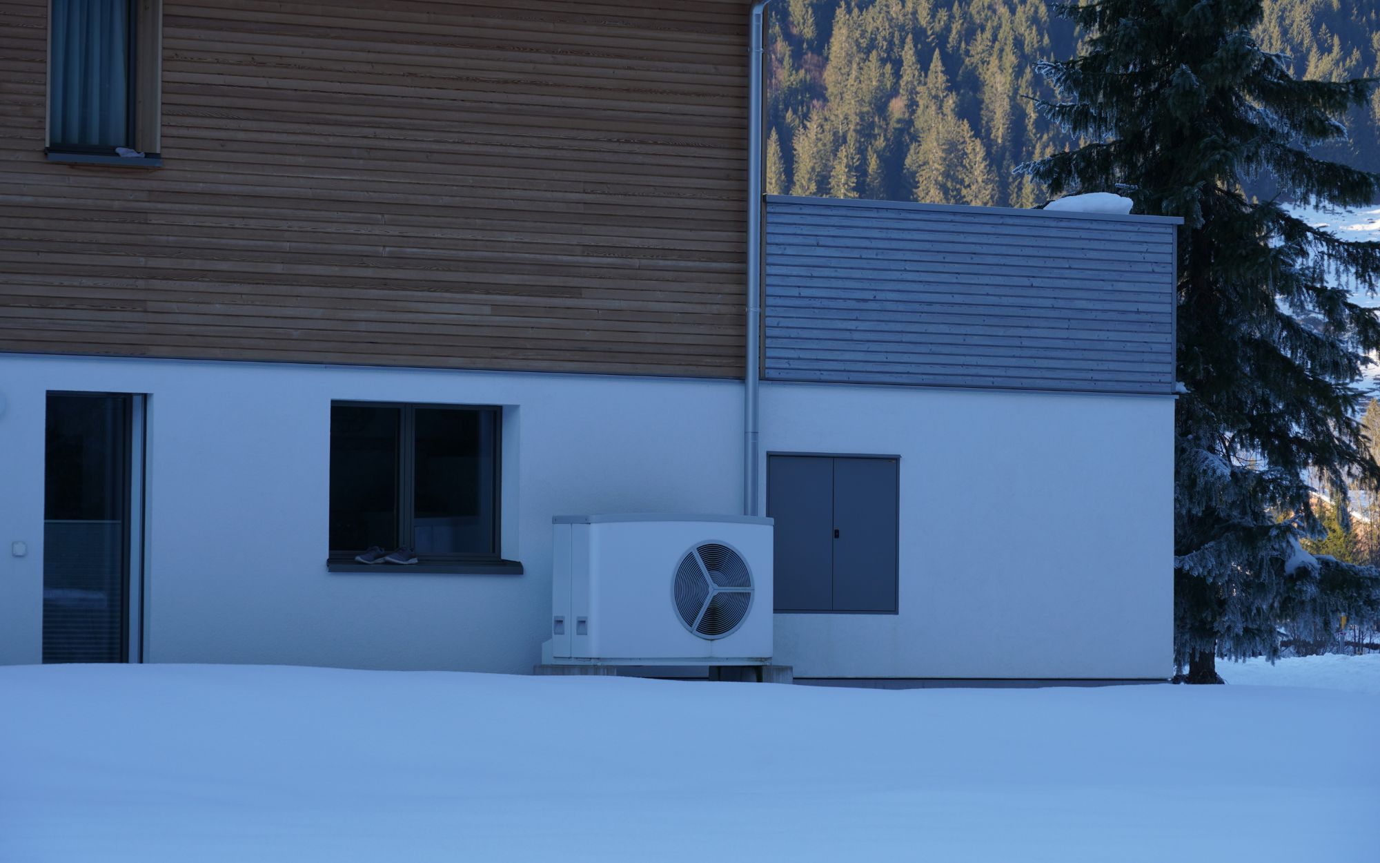 Outdoor heat pump unit installed beside a modern home surrounded by snow, with evergreen trees and mountains in the background.