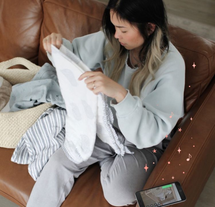 A woman folds laundry on a couch while her phone shows a view of her baby sleeping.