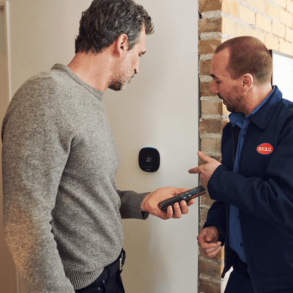 An HVAC technician points to an ecobee thermostat while a customer checks the ecobee mobile app